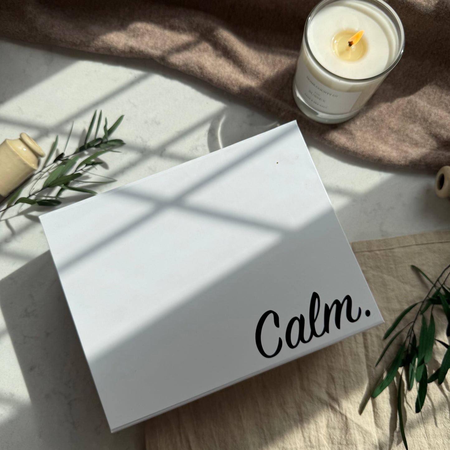 White box with 'Calm.' text on a surface with a candle and plants