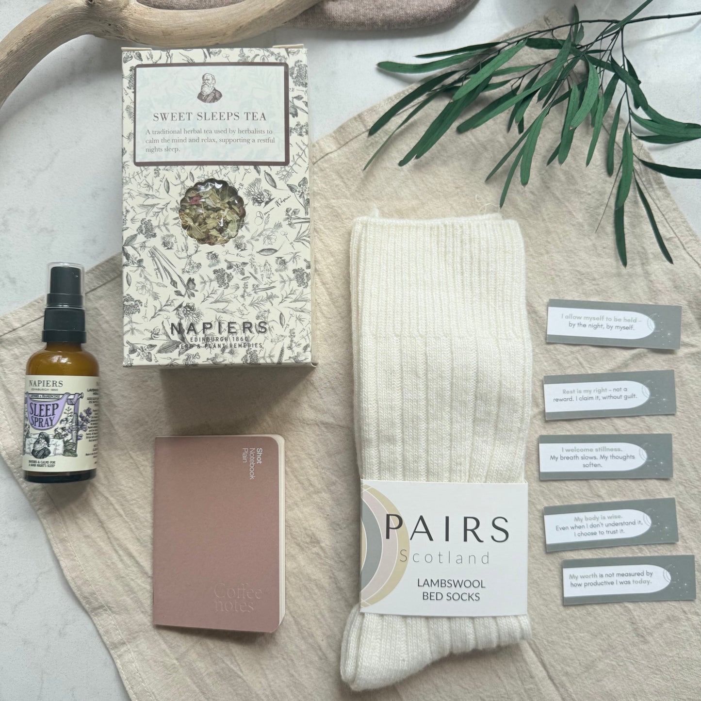 Set of items including a bottle, box, notebook, and socks on a textured surface with greenery.