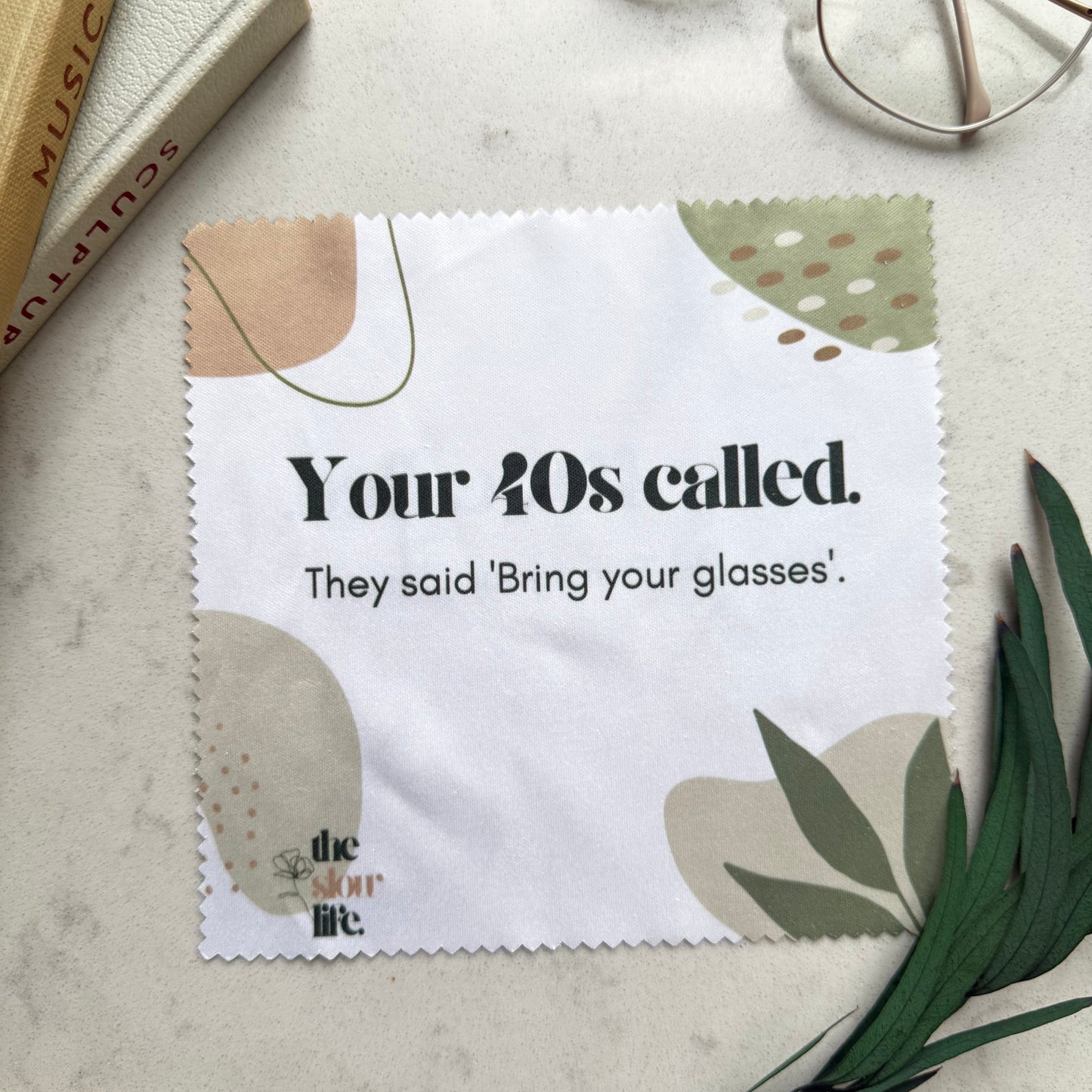 Cleaning cloth with text 'Your 40s called. They said 'Bring your glasses'.', placed on a surface with books and glasses.