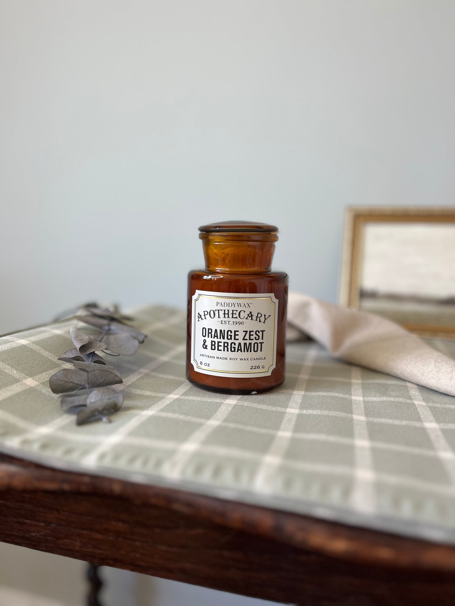 Amber glass jar candle labeled 'Apothecary Glass Jar Candle - Orange Zest & Bergamot', with a vintage-inspired design and a warm, inviting glow.