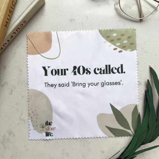 Cleaning cloth with text 'Your 40s called. They said 'Bring your glasses'.', placed on a surface with books and glasses.