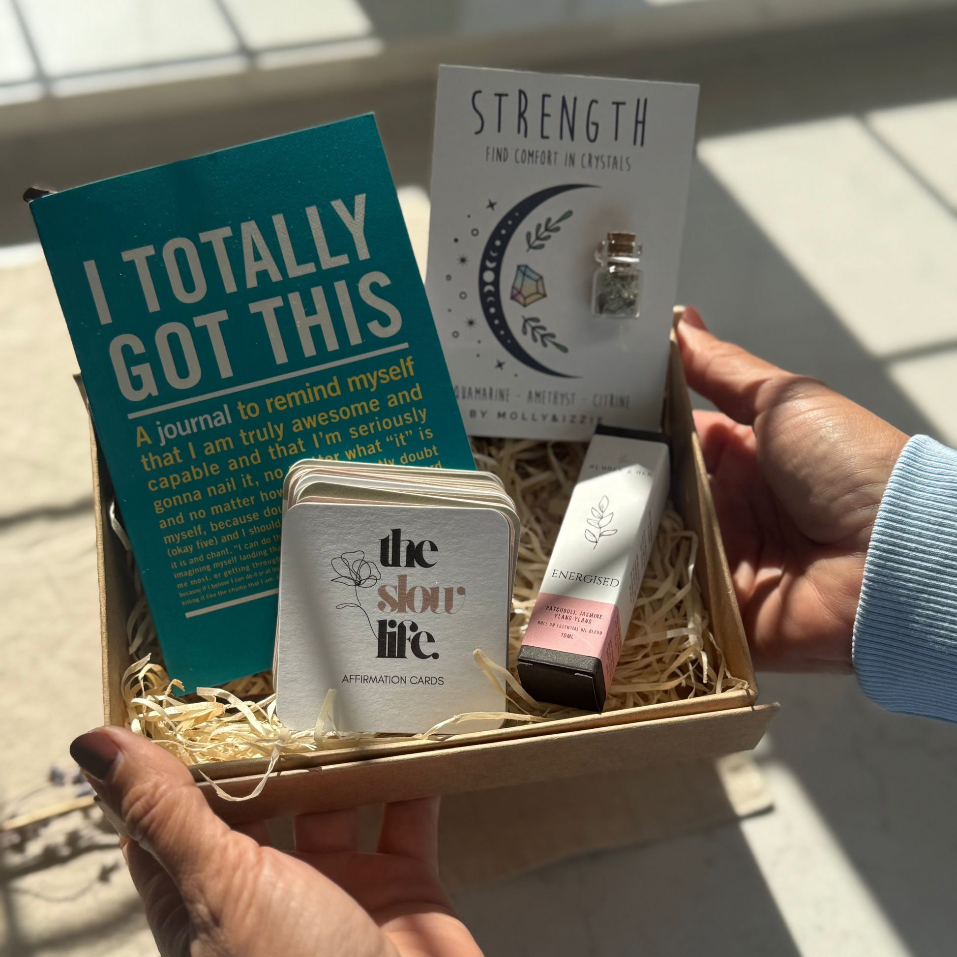 Hand holding a gift box with 'I Totally Got This' journal, 'Strength' card, and other items.