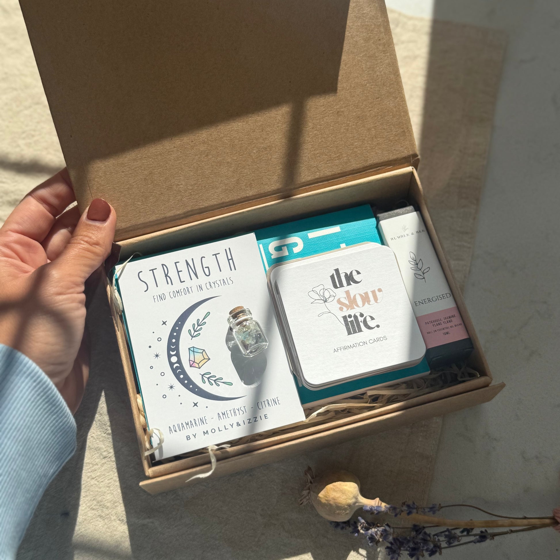 Hand opening a box containing cards and a small bottle labeled 'the slow life' on a neutral background.