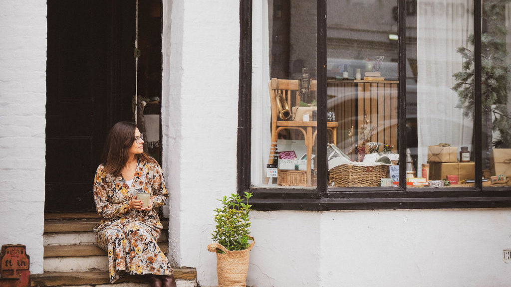 Woman sitting on a step outside a white shop with a window display.